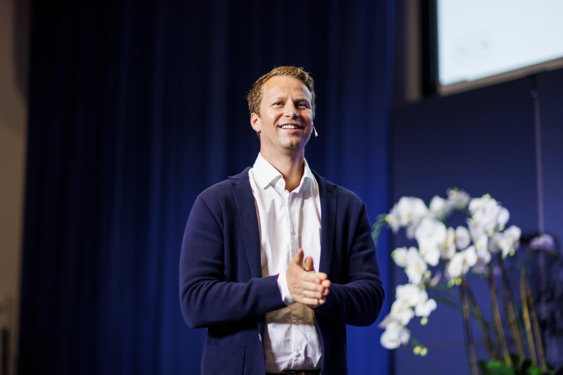 Mads Nordmo Arnestad på scenen under et foredrag, smilende, med sceneteppe og blomster i bakgrunnen